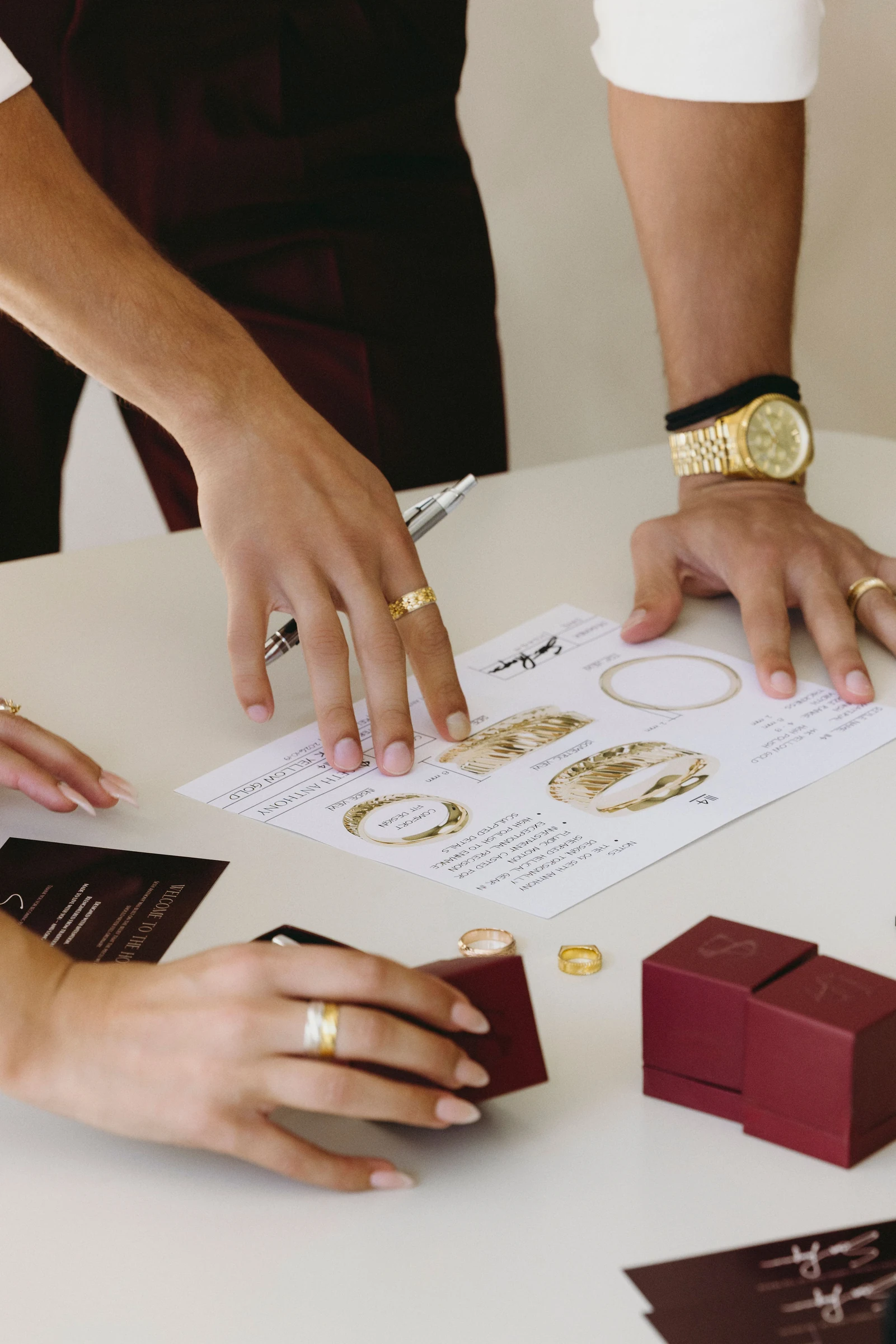 Hands arranging burgundy ring boxes around a ring design drawing