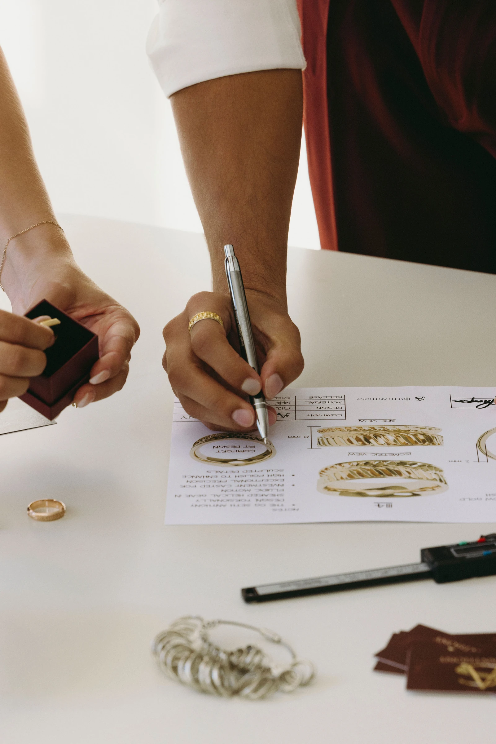 A hand marking a ring design drawing with a pen beside calipers and ring boxes